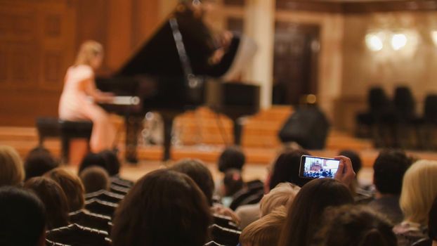 Spectators in concert hall during performing piano girl- people shooting performance on smartphone, telephoto