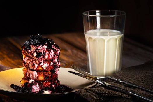 Fried cheesecakes with blueberry jam and sour cream on a plate and a glass of milk on the table.