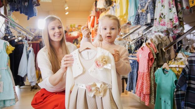 Blonde young woman with little daughter buying kids dress in clothes store, telephoto
