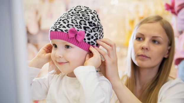 Little blonde girl with mommy doing shopping and buying hat in kids dress store, telephoto