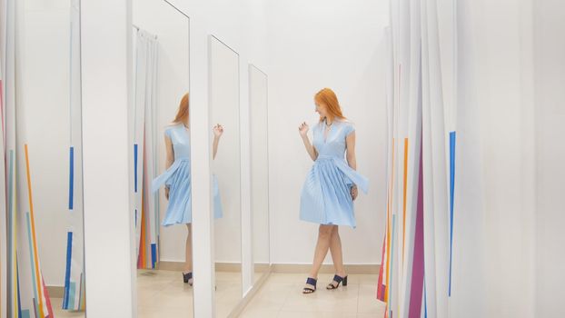 Young woman trying blue dress near mirror in fitting room at store, middle shot