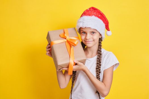 emotional cute teenager brown-hair girl in santa claus hat with a gift on a yellow background, christmas gifts concept copy space