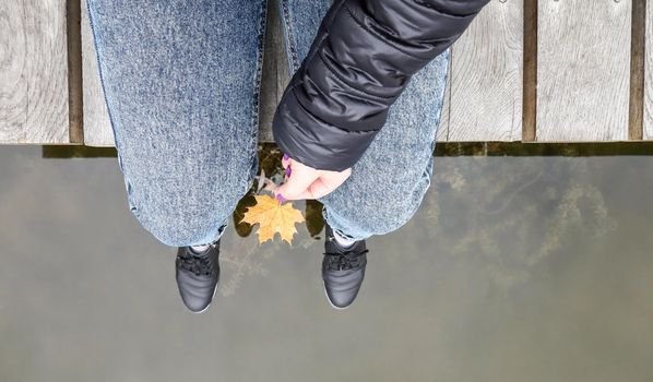 Beautiful legs of a girl in jeans are sitting on a wooden bridge by the lake, holding a yellow maple leaf in their hands. Autumn warm and sunny day. View from above. Autumn concept.
