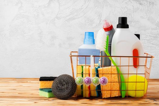 Cleaning tools and chemicals in basket on wooden table against gray background, front view