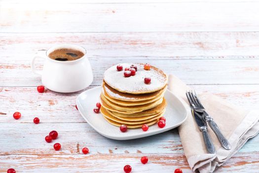 morning breakfast of pancakes with cranberries and powdered sugar on a wooden table and a cup of coffee.