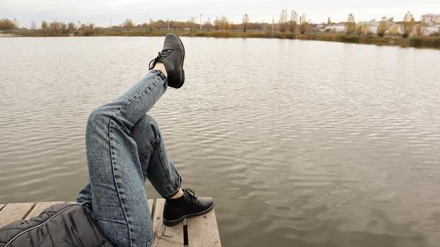 A girl in blue jeans sits by the lake. A young woman is resting by the river, sitting on the edge of a wooden bridge. Pair of legs relaxing by a lake on a wooden dock. People are resting.