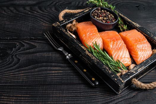 Raw salmon fillet fish steaks in a wooden tray with thyme and rosemary. Black wooden background. Top view. Copy space.