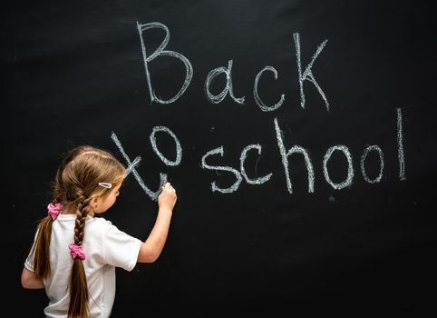 little girl in a white blouse with pigtails writes with chalk on black chalkboard back to school