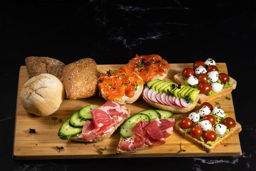 An assortment of sandwiches with fish, cheese, meat and vegetables lay on the board and a bun.