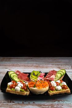 Assorted sandwiches with fish, cheese, meat and vegetables on a black plate and wooden background.