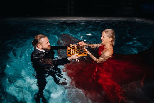 a man in a suit and a girl in a red dress play chess on the water in the pool.