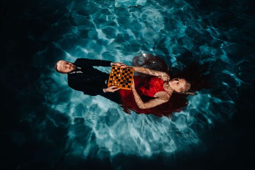 a man in a suit and a girl in a red dress are lying on the water with chess in the pool.