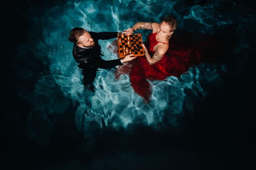 a man in a suit and a girl in a red dress play chess on the water in the pool.