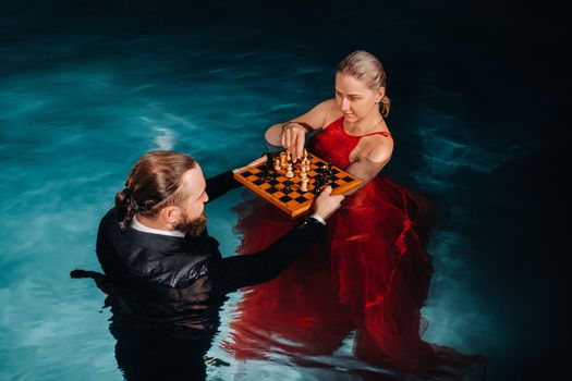 a man in a suit and a girl in a red dress play chess on the water in the pool.