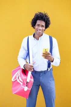 Black man with afro hairstyle carrying a sports bag and smartphone against a yellow urban background. Guy with curly hair wearing shirt and suspenders.