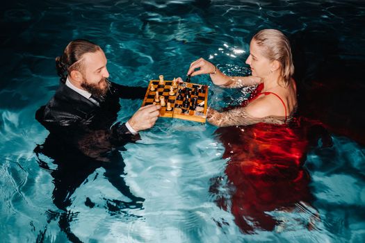 a man in a suit and a girl in a red dress play chess on the water in the pool.