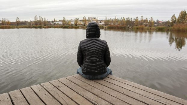 A girl in a jacket sits on a wooden bridge by the lake in cold autumn weather. Dipresia or dreaming alone. Thinking one on one with nature