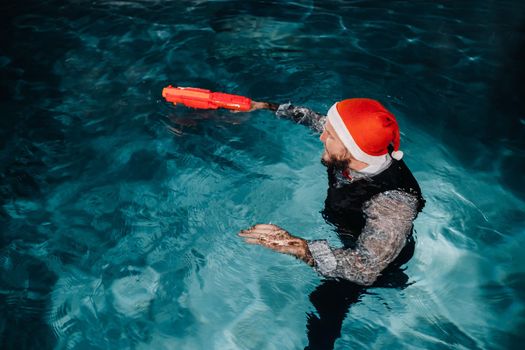 A man in a Santa Claus suit and hat with a red gun in his hands. Santa Claus is swimming underwater with a gun.Christmas concept