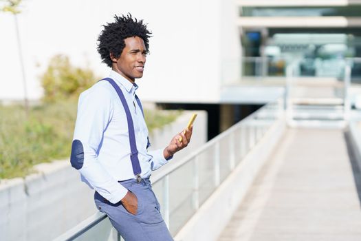 Black man with afro hairstyle using a smartphone near an office building. Guy with curly hair wearing shirt and suspenders.