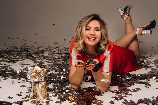 A girl in a red dress is lying on the floor with a bunch of gifts on a gray background in confetti.