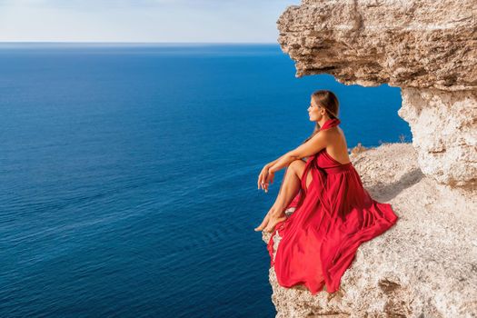 Side view a Young beautiful sensual woman in a red long dress posing on a rock high above the sea during sunrise. Girl on the nature on blue sky background. Fashion photo.