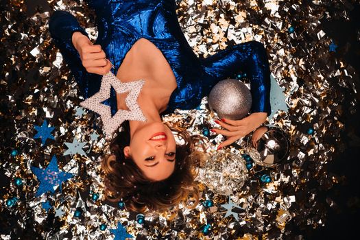 a woman in a blue sequined dress smiles and lies on the floor under a falling multicolored confetti.