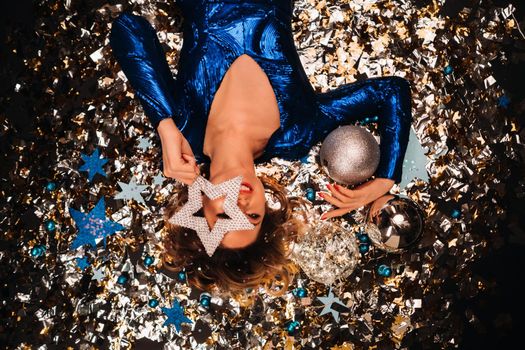 a woman in a blue sequined dress smiles and lies on the floor under a falling multicolored confetti.
