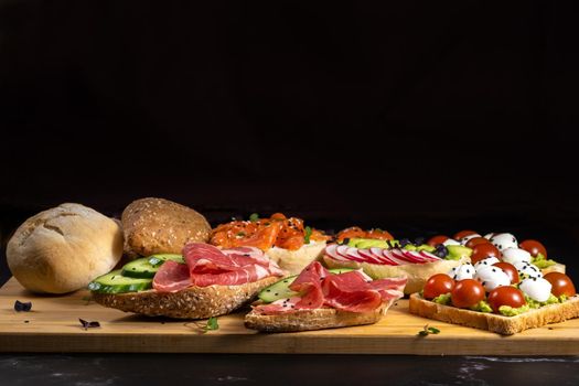 An assortment of sandwiches with fish, cheese, meat and vegetables lay on the board and a bun.