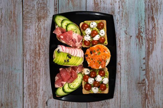 Assorted sandwiches with fish, cheese, meat and vegetables on a black plate and wooden background.
