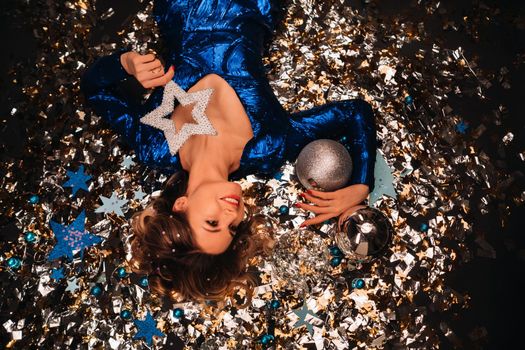 a woman in a blue sequined dress smiles and lies on the floor under a falling multicolored confetti.