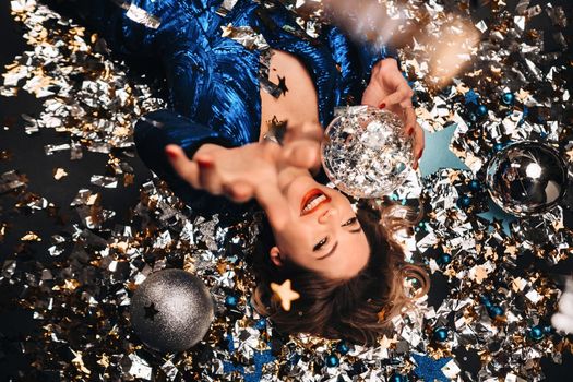 a woman in a blue sequined dress smiles and lies on the floor under a falling multicolored confetti.