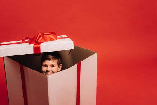 A little boy sits in a Christmas present and looks out of it on a red background.