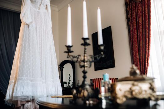 Vintage wedding dress hanging on a wooden hanger and a candle holder standing on a table in the castle.