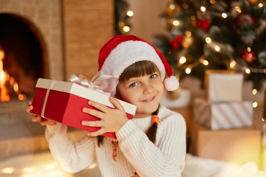 Funny child in Santa red hat holding Christmas gift in hand and looking directly at camera, trying to guess what is in present box, sitting in living room near fireplace and x-mas tree.