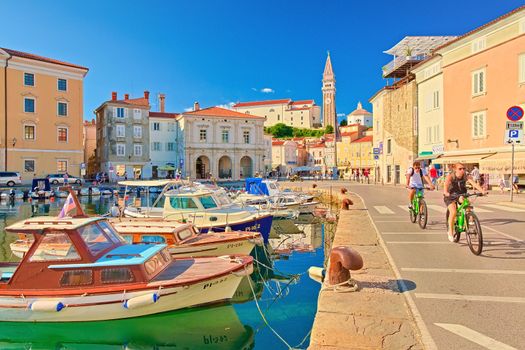 Piran - July 2020, Slovenia: A couple ride bicycles on the pier near colorful boats lying on the water in the harbor of the ancient Slovenian town of Piran