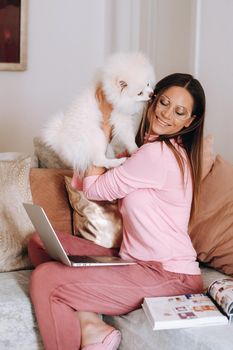 a girl in pajamas at home is working on a laptop with her dog Spitzer, the dog and its owner are resting on the couch and watching the laptop.Household chores.