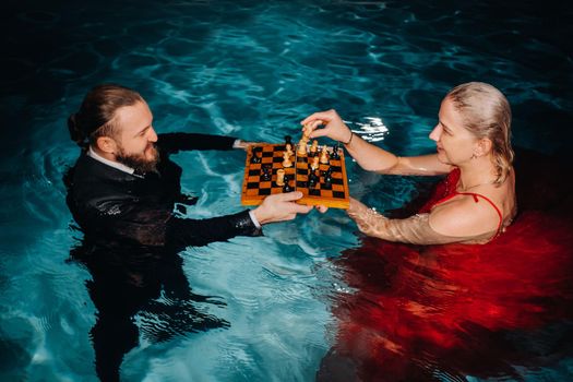 a man in a suit and a girl in a red dress play chess on the water in the pool.