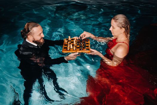 a man in a suit and a girl in a red dress play chess on the water in the pool.