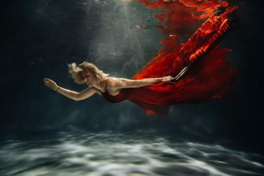 A girl in a red dress is floating under the water with a suitcase.Journey under the water of a single woman.The concept of underwater tourism.
