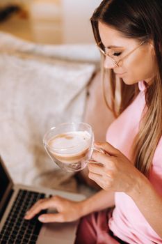a girl in the morning in pajamas at home working on a laptop with drinking coffee, a girl self-isolated at home and resting on the couch and watching a laptop.Household chores.