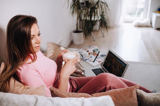 a girl in the morning in pajamas at home working on a laptop with drinking coffee, a girl self-isolated at home and resting on the couch and watching a laptop.Household chores.