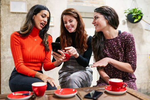 Multiethnic group of three happy female friends looking to a smartphone in a cafe bar.