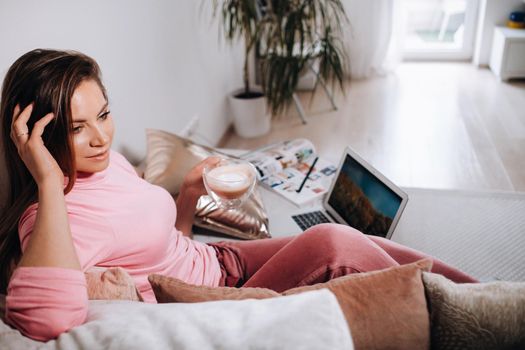 a girl in the morning in pajamas at home working on a laptop with drinking coffee, a girl self-isolated at home and resting on the couch and watching a laptop.Household chores.