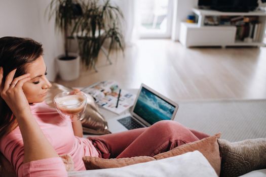 a girl in the morning in pajamas at home working on a laptop with drinking coffee, a girl self-isolated at home and resting on the couch and watching a laptop.Household chores.