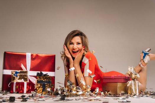 A girl in a red dress is lying on the floor with a lot of gifts on a gray background.