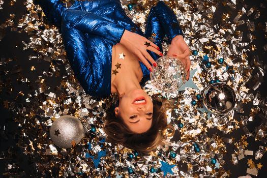 a woman in a blue sequined dress smiles and lies on the floor under a falling multicolored confetti.