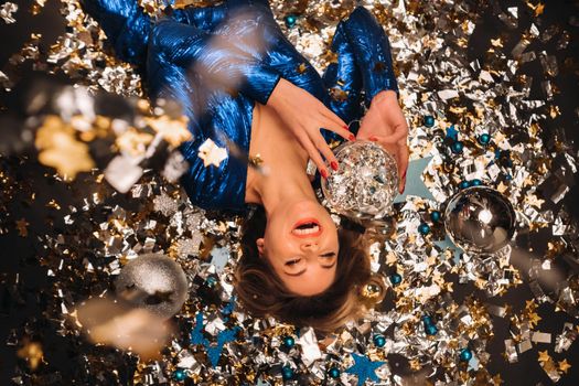 a woman in a blue sequined dress smiles and lies on the floor under a falling multicolored confetti.