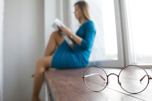 Beautiful young blonde female without glasses sitting near window and reading book, telephoto