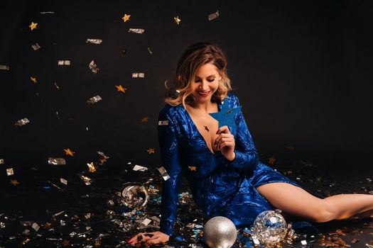 a woman in a blue sequined dress is smiling sitting on the floor with confetti on a black background.