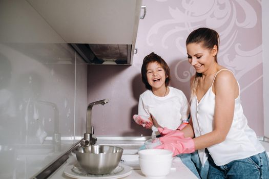 housewife mom in pink gloves washes dishes with her son by hand in the sink with detergent. A girl in white and a child with a cast cleans the house and washes dishes in homemade pink gloves.A child with a cast washes dishes and smiles.